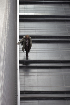 elderly woman with clear jacket and dark pants coming down stairs with a wide black tiles and metal...