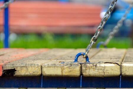 A gray chain with the remains of yellow paint holds a hinged bridge on a playground with a blurred...