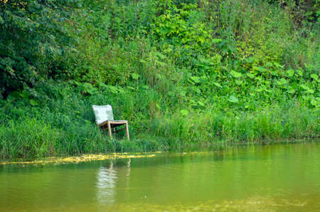 An old wooden chair stands on the shore of a pond with dirty waterの写真素材