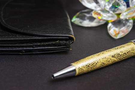 An old black leather wallet with traces of scuffs and stitching. Next to it is a pen for writing and a glass product.の写真素材