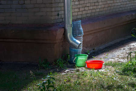A bucket, once and other means of collecting water stand near the tin drain pipe. Next to the brick...