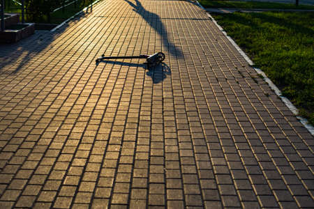 A small scooter lies on the sidewalk in the setting sun. Then the shadow of man.