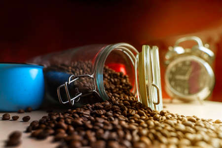 Coffee beans from a glass jar are poured onto the table. Near the clock and mug