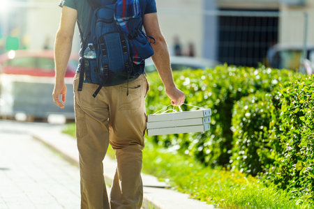 View from the back. A man with a backpack carries three boxes of pizza in the direction of the sun in the city.の写真素材