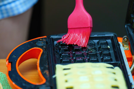 view of a pink silicone brush spreading oil on an electric waffle maker, highlighting the process of...