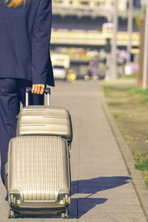 young woman in a blue suit confidently rolls her suitcases along the sidewalk, heading towards the road. Her poised demeanor suggests a journey ahead, blending style with purposeの写真素材