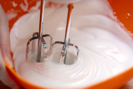 metal mixer beating a translucent egg white into a stiff white foam, set against a vibrant orange bowl, showcasing the process of baking and food preparationの写真素材