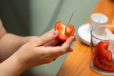 hand cuts a red tomato into wedges with a knife, preparing fresh ingredients for blending in a culinary recipe.の写真素材