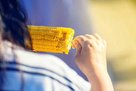 Back view of a girl eating boiled corn on a wooden stick with salt on a bright sunny day. Casual...