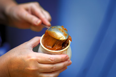 Girl's hand scooping thick honey with metal spoon into ceramic pot for sale.