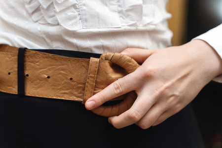 A girl's hand adjusting a vintage wide beige leather belt with coiled leather buckle.の写真素材