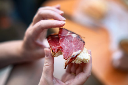A bitten sandwich with bread and cured red meat in female hands during breakfast, ideal for food blogs and lifestyle content.の写真素材