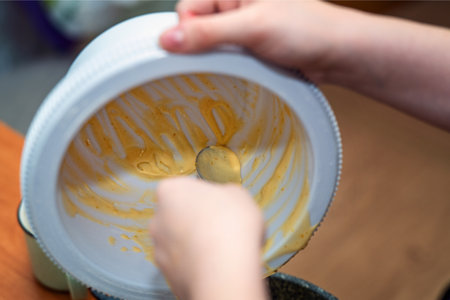 Female hand scraping leftover batter from stand mixer bowl with metal spoon, illustrating frugality...