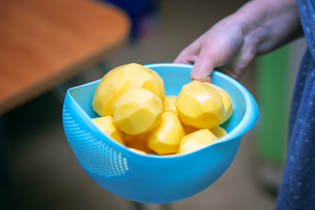 Woman's hand carrying a blue plastic bowl filled with peeled potatoes.の写真素材