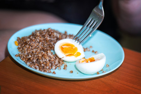 Boiled chicken egg halved by metal fork on blue ceramic plate with buckwheat porridge.の写真素材