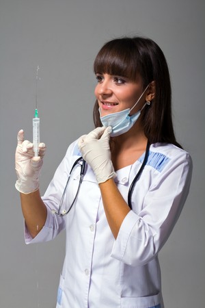 Smiling young doctor with mask, stethoscope and syringe on a grey backgroundの写真素材