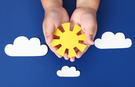 Child holds in his palms a paper sun in the blue sky with paper clouds.の写真素材