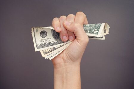 Woman hand squeezes dollar bills against grey background.