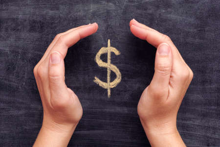 Hands protecting drawn dollar sign on black chalkboard background. Close up.の写真素材