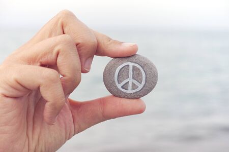 Person holds stone with peace symbol against sea in background. Close up.の写真素材