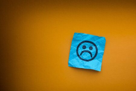 Blue paper note with sad face on a yellow paper background. Depression concept.の写真素材