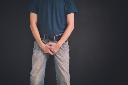 Man in jeans covers his crotch with hands against black wall. Close up.