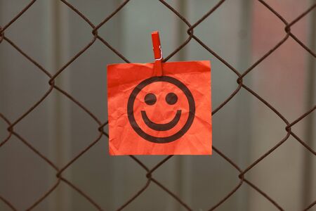 Paper note with happy face hanging on a metal mesh fence. Close up.の写真素材