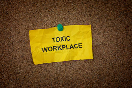A crumpled sticky note with the words Toxic Workplace on it pinned to a cork board. close up.の写真素材