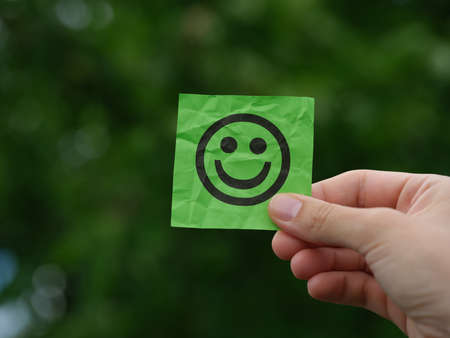 A woman holding a green sticky note with a happy face on it in her hand against a background of foliage. close up.の写真素材