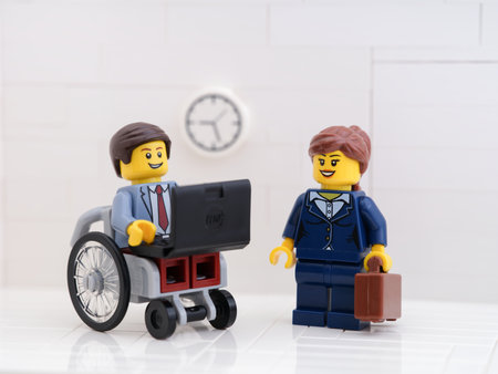 Tambov, Russian Federation - September 6, 2022 A Lego businessman minifigure in a wheelchair using a laptop with a lego businesswoman minifigure standing next to him and holding a briefcase.のeditorial素材