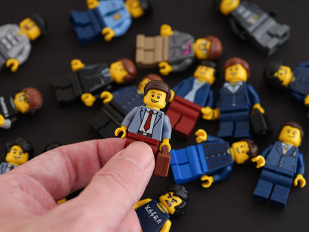 Tambov, Russian Federation - October 09, 2022 A hand holding a Lego businessman minifigure against a pile of minifigure, which are lying on a black background.のeditorial素材