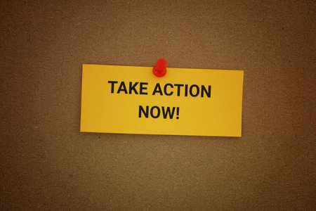 A yellow paper note with the phrase Take Action Now! on it pinned to a cardboard background. Close up.の写真素材