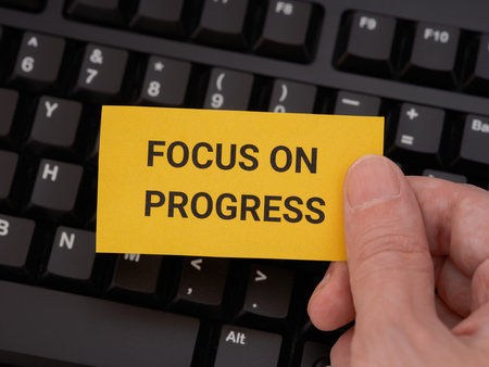 A man holding a yellow paper note with the phrase Focus On Progress on it in his hand against a black computer keyboard. Close up.の写真素材