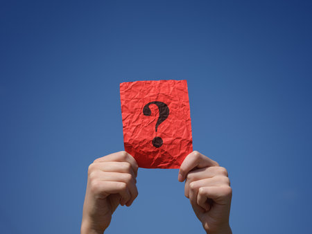 A boy holding a crumpled red paper note with a question mark on it in his hands against the blue sky. Close up.の写真素材