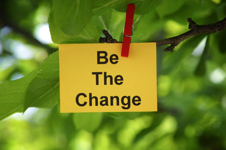 A yellow paper note with the phrase Be The Change on it attached to a tree branch with a clothes pin. Close up.の写真素材