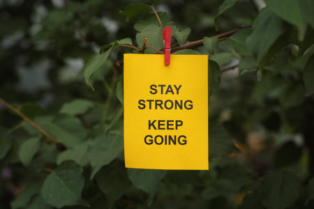 A yellow paper note with the words Stay Strong Keep Going on it attached to a tree branch with a clothes pin. Close up.の写真素材