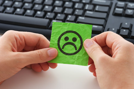 A businesswoman in an office sitting behind a computer and holding a green piece of paper with a sad face on it in her hands. Stress at the workplace. Close up.の写真素材