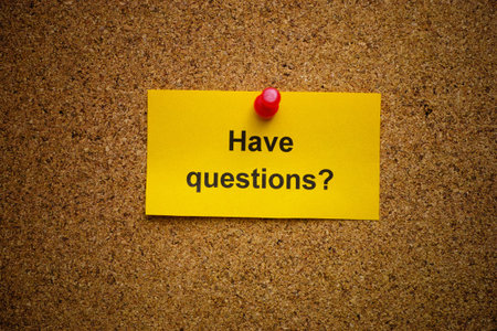 A yellow paper note with the question Have questions on it pinned to a cork board. Close up.の写真素材