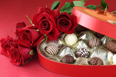 Heart shaped box filled with a variety of candies and long stem roses against a red background. Selective focus on candy.の写真素材