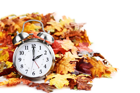 Alarm clock in colorful autumn leaves isolated against a white background with light shadow and shallow depth of field. Daylight savings time concept.の写真素材
