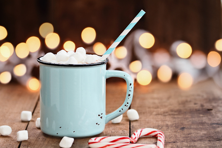 Enamel cup of hot cocoa with mini marshmallows and candy canes in the shape of a heart against a rustic background with beautiful Christmas lights of bokeh. Could also be coffee. Perfect winter time treat.の写真素材