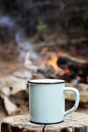 Blue enamel cup of hot steaming coffee sitting on an old log by an outdoor campfire. Extreme shallow depth of field with selective focus on mug.の写真素材