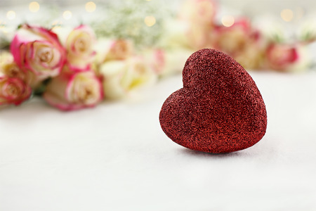 A red glitter heart in front of long stem white and red roses and babys breath with fairy lights in the background. Shallow depth of field with selective focus on foreground.の写真素材