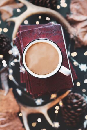 Hot, steaming cup of coffee over a stack of books with dried autumn leaves, deer antlers, pines cones, and sparkling lights. Selective focus on mug with extreme blurred background. Top view. の写真素材