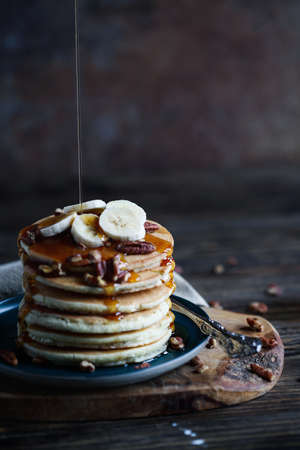 Maple syrup pouring onto pancakes served with fresh banana fruit and nuts. Selective focus with...