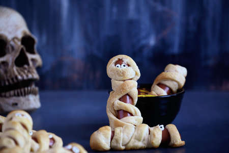 Fun food for kids. Mummy hot dogs on a blue rustic table with a bowl of ketchup and mustard dip,...
