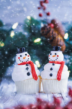 Two iced Christmas Snowman cupcakes with carrot nose, santa hat, and scarf. Selective focus with blurred foreground and background.の写真素材