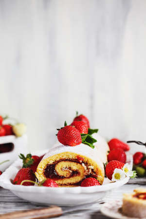 Homemade strawberry shortcake cake roll, Sponge or Roulade with a berry jam filling and powdered sugar with mint leaves. Dessert over a white rustic wooden table. Selective focus with blurred foreground and background.の写真素材
