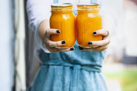 Woman in an apron holding out fresh homemade peach butter. Home preservation. Selective focus on...