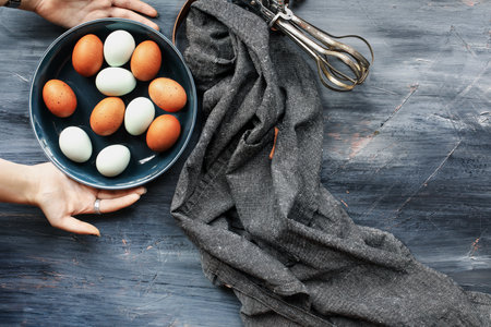 Young woman's hands placing a bowl of fresh brown and blue eggs onto a rustic wooden table. Top view.の写真素材